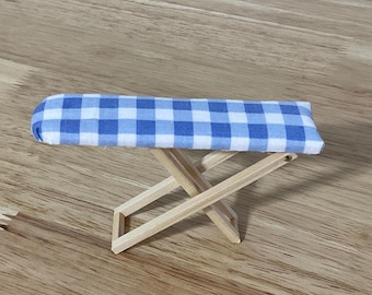 Puede incluir: Una tabla de planchar en miniatura con una funda de tela de cuadros vichy azul y blanco sobre una superficie rectangular, sostenida por un marco plegable de madera clara con diseño de patas en cruz y pivotes metálicos.