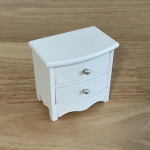 A small white wooden nightstand with two drawers is placed on a wooden surface.