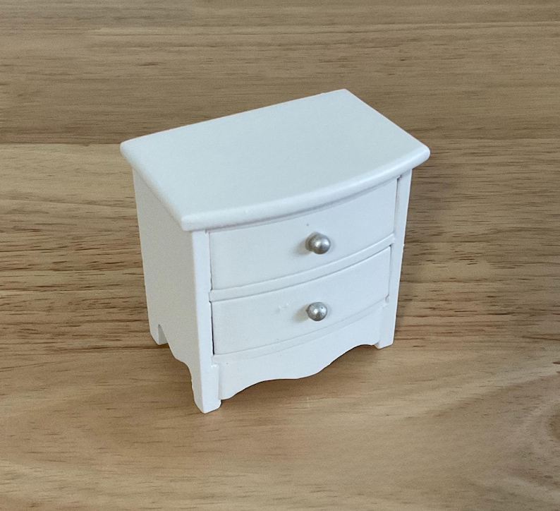 A small white wooden nightstand with two drawers is placed on a wooden surface.