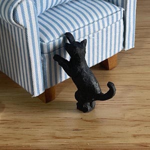 May include: A miniature black cat figurine standing on its hind legs with its front paws resting against the side of a small armchair upholstered in a blue and white striped fabric with light brown wooden legs.