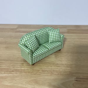 May include: A miniature sofa upholstered in a green and white gingham check fabric. The piece features two matching throw pillows, a structured backrest, and small brown wooden feet, designed with a classic silhouette and clean lines.