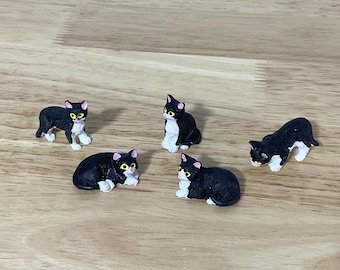 May include: A set of four miniature cat figurines, each featuring a black and white coat, pink ears, and bright yellow eyes. Two of the cats are depicted in a standing position, while the other two are shown in a resting, lying-down pose.
