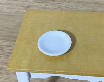 May include: A small, round, white plate sits centered on a flat, mustard-yellow surface. The plate has a smooth, matte finish and a slightly raised rim, appearing as a miniature item for a dollhouse or a small-scale display setting.