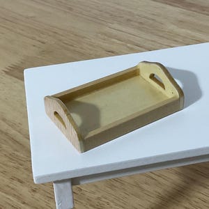 May include: A small, rectangular wooden tray with a light yellow interior and natural wood-colored frame. The tray has curved handles on each end. It sits on a white table, with a wooden floor in the background. The tray is likely for serving or display.