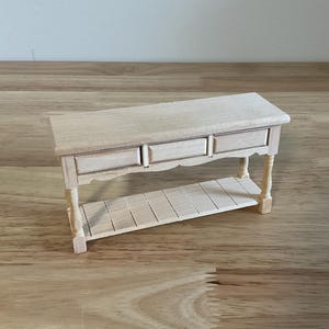 May include: A miniature wooden console table featuring a rectangular top, three small drawers across the front, and a lower slatted shelf. The piece is constructed from light-colored, unfinished wood and stands on four slender, turned legs.