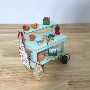 May include: A miniature light blue wooden potting bench featuring two shelves, decorated with tiny terracotta pots, a red and green birdhouse, small gardening tools, a floral apron, a bag labeled Organic Potting Mix, and seed packets.