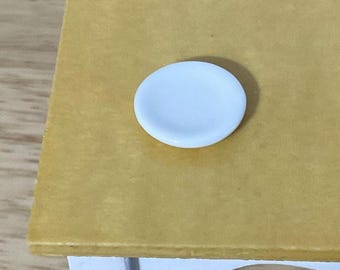 May include: A small, round, white plate sits on a flat, mustard-yellow surface. The plate has a smooth, solid white finish with a slightly raised edge, appearing as a miniature item placed on a larger, rectangular yellow tabletop.