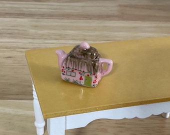 May include: A miniature ceramic teapot shaped like a cottage with a pink exterior, a brown thatched-style roof, a green door, and small painted red flowers. The teapot features a spout on the left, a handle on the right, and a small lid on top.