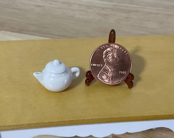 May include: A miniature white ceramic teapot with a lid, handle, and spout, positioned next to a 2015 United States copper penny held upright on a small brown wooden display stand to show the scale of the tiny object.