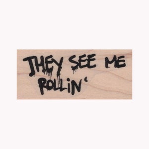May include: A wooden stamp with the phrase "THEY SEE ME ROLLIN'" in black, dripping font. The stamp has a light wood grain background.