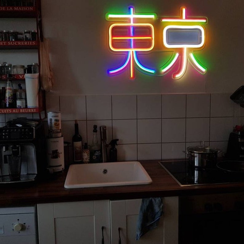 May include: A rainbow neon sign with the Chinese characters for "Tokyo" on a white tiled wall in a kitchen. The sign is lit up and casting a colorful glow on the surrounding area.