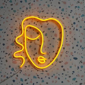 May include: A neon sign in the shape of a face with a wavy line for hair. The sign is lit up in a bright yellow color and is mounted on a white wall with a speckled pattern.