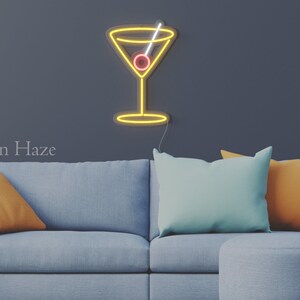 May include: A yellow neon sign shaped like a martini glass with a white straw and a red olive. The sign is mounted on a dark wall above a light blue sofa with two orange pillows and one light blue pillow. The text Neon Haze is visible.