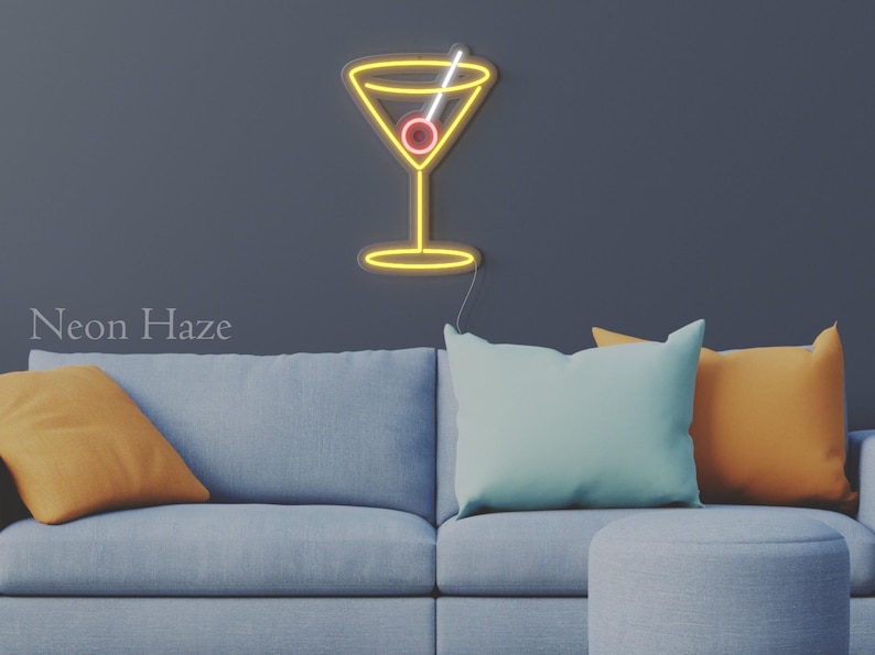 May include: A yellow neon sign shaped like a martini glass with a white straw and a red olive. The sign is mounted on a dark wall above a light blue sofa with two orange pillows and one light blue pillow. The text Neon Haze is visible.