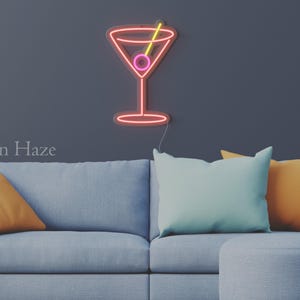 May include: A wall-mounted neon sign shaped like a martini glass with a pink olive and a yellow toothpick. The sign features red and pink neon tubing and is mounted on a dark wall above a gray sofa with orange and light blue throw pillows. Text reads Neon Haze.