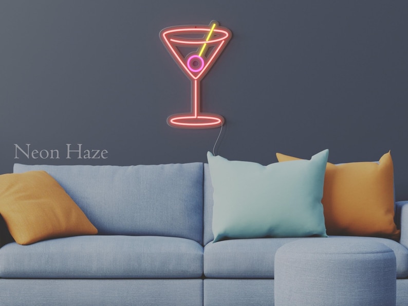 May include: A wall-mounted neon sign shaped like a martini glass with a pink olive and a yellow toothpick. The sign features red and pink neon tubing and is mounted on a dark wall above a gray sofa with orange and light blue throw pillows. Text reads Neon Haze.