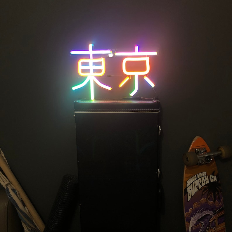 May include: A neon sign with the word "Tokyo" in Japanese characters. The sign is lit up in a rainbow of colors and is mounted on a black amplifier case.