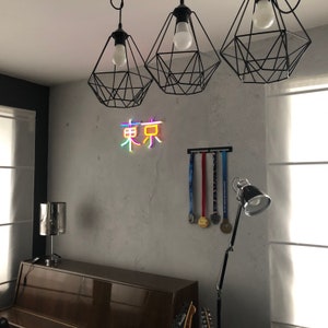 May include: A room with a gray wall, a piano, and a neon sign that says "Tokyo" in Japanese characters. There are three black wire cage light fixtures hanging from the ceiling. A black metal rack with medals hanging on it is on the wall.