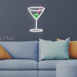 May include: A wall-mounted neon sign shaped like a martini glass with a white outline, a green olive, and a pink toothpick. The sign features a thin power cord extending from the bottom and is displayed against a dark gray wall above a sofa.