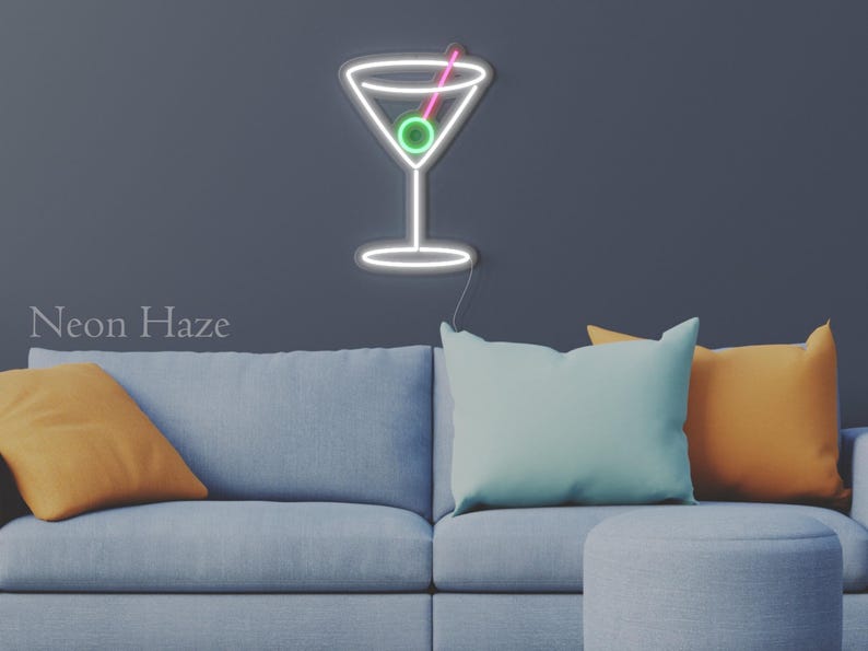 May include: A wall-mounted neon sign shaped like a martini glass with a white outline, a green olive, and a pink toothpick. The sign features a thin power cord extending from the bottom and is displayed against a dark gray wall above a sofa.
