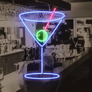 May include: A neon sign shaped like a martini glass with a blue outline, a bright green olive in the center, and a pink toothpick. The sign is displayed against a dark, blurred background of a bar setting with glassware and bottles.