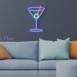May include: A wall-mounted neon sign shaped like a martini glass with a blue outline, a green olive, and a pink toothpick. The sign is illuminated against a dark gray wall above a light gray sofa with orange and light blue throw pillows.