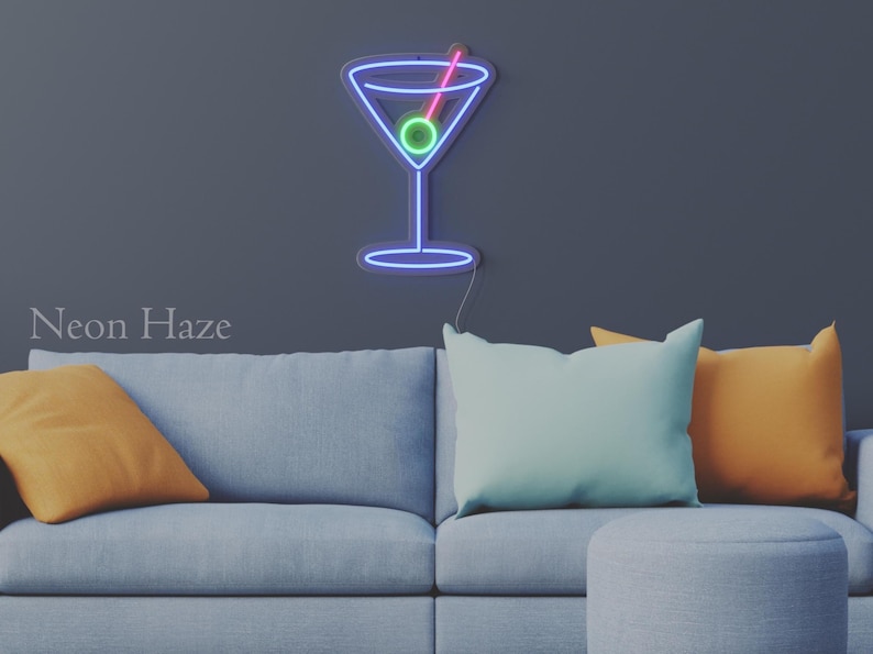 May include: A wall-mounted neon sign shaped like a martini glass with a blue outline, a green olive, and a pink toothpick. The sign is illuminated against a dark gray wall above a light gray sofa with orange and light blue throw pillows.