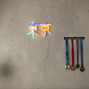 May include: A neon sign with the Japanese characters for "Tokyo" in pink, yellow, and blue. Below the sign, a black metal rack holds four medals with red, blue, and gold ribbons.