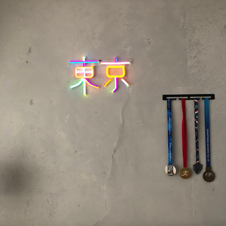 May include: A neon sign with the Japanese characters for "Tokyo" in pink, yellow, and blue. Below the sign, a black metal rack holds four medals with red, blue, and gold ribbons.