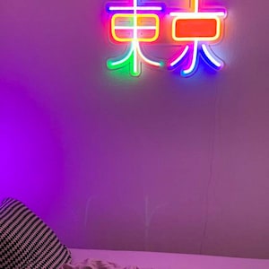 May include: A neon sign with the Chinese character for "East" in rainbow colors, mounted on a purple wall. The sign is lit up and casting a glow on the wall.
