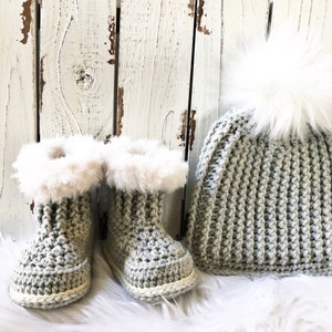 May include: A set of gray crocheted baby booties with white faux fur trim and a matching gray crocheted beanie with a white pom pom.