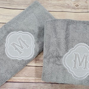 May include: Two gray towels with white embroidered monograms in the shape of a decorative frame. The monogram is the letter "M".