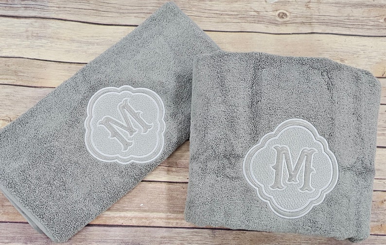 May include: Two gray towels with white embroidered monograms in the shape of a decorative frame. The monogram is the letter "M".