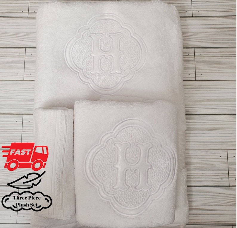 May include: Three white plush towels with an embroidered letter "H" in a decorative circle design. The towels are arranged in a stack, with the largest towel on the bottom and the smallest towel on top.