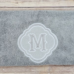 May include: A gray bath mat with a white embroidered monogram of the letter "M" in a decorative frame.