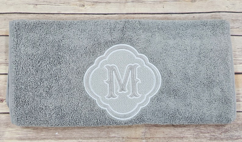 May include: A gray bath mat with a white embroidered monogram of the letter "M" in a decorative frame.
