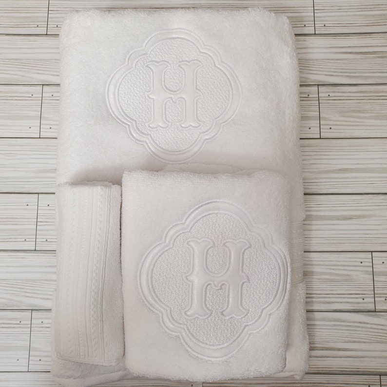 May include: Three white towels with embroidered letter H in a decorative frame. The towels are folded and stacked on a white wooden surface.