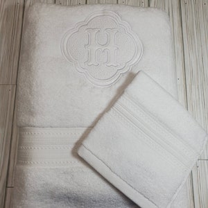 May include: Two white bath towels, one folded on top of the other. The top towel has a monogrammed "H" in a decorative frame.