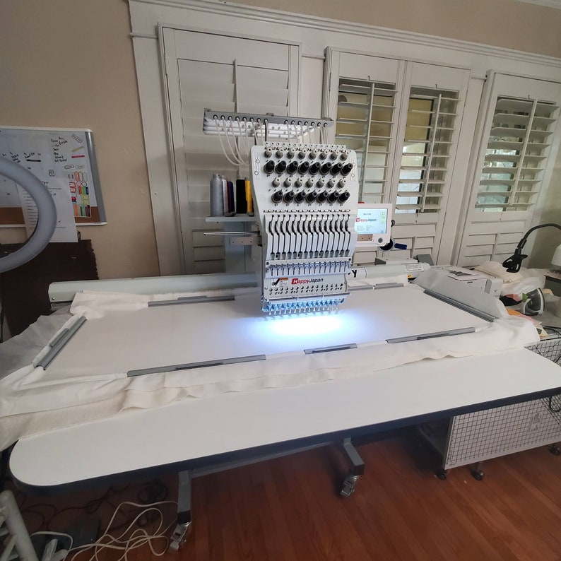 May include: A white industrial embroidery machine with a large needle head and a white table with a white fabric stretched across it. The machine is in a room with white walls and windows with white blinds.