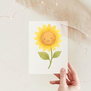 Könnte beinhalten: Ein rechteckiger Druck mit einer Aquarellillustration einer leuchtend gelben Sonnenblume mit lächelndem Gesicht, rosigen Wangen und brauner Mitte. Die Blume hat einen grünen Stängel und zwei grüne Blätter vor einem schlichten weißen Hintergrund.