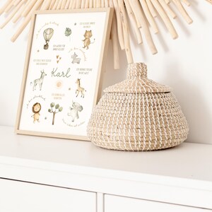 May include: A framed print with watercolor illustrations of animals and a hot air balloon, with the name "Karl" in the center. A woven basket with a lid sits beside the frame. A decorative mirror with wooden accents is in the background.