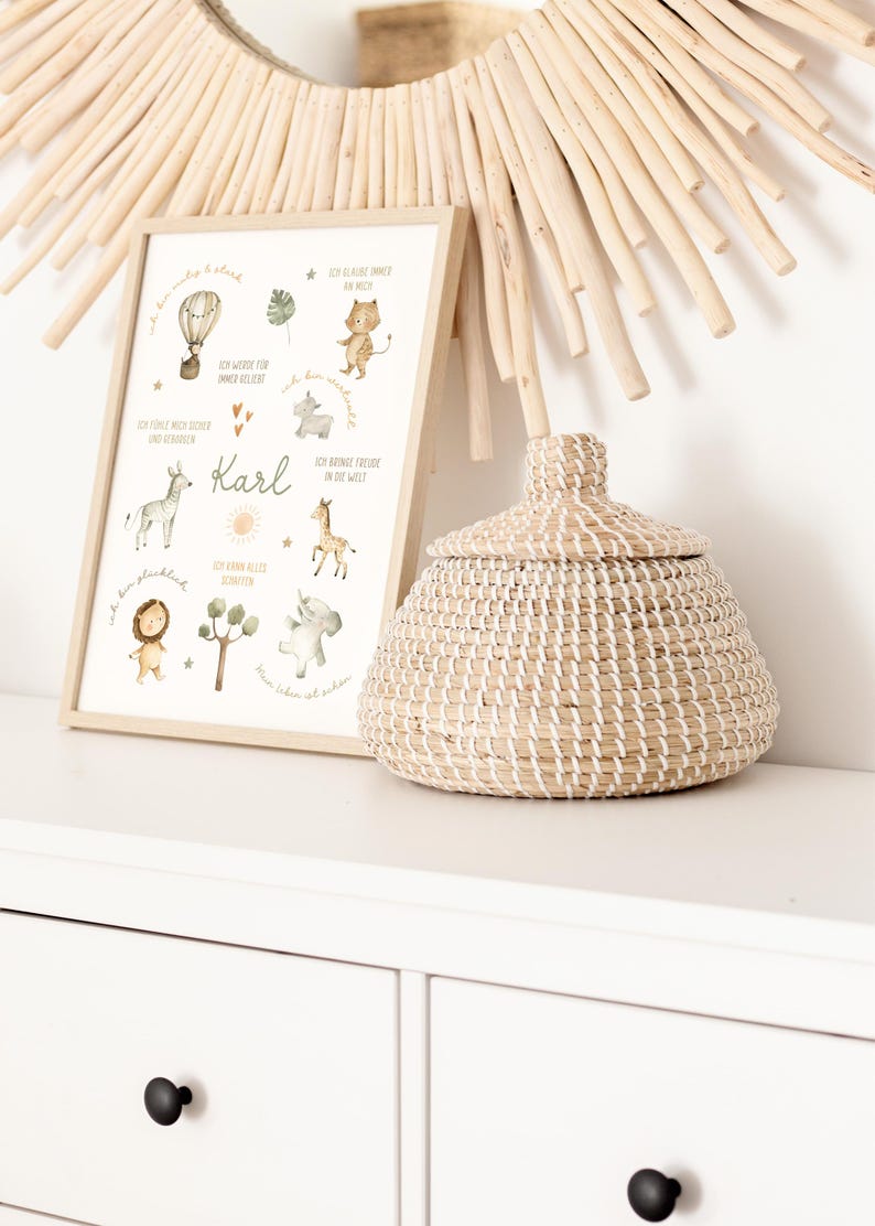 May include: A framed print with watercolor illustrations of animals and a hot air balloon, with the name "Karl" in the center. A woven basket with a lid sits beside the frame. A decorative mirror with wooden accents is in the background.