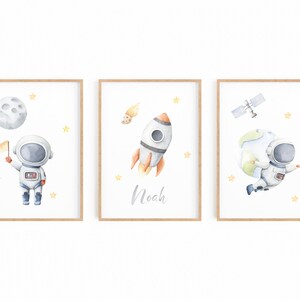 以下が含まれることがあります： 宇宙をテーマにしたイラストが描かれた、額入りの水彩画プリント3枚セットです。月面に着陸した宇宙飛行士、名前「Noah」のロケット、地球を持つ宇宙飛行士が描かれています。各プリントは木製のフレームで、黄色の星のアクセントが付いています。