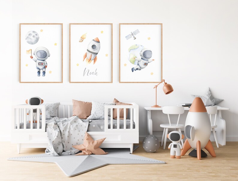 以下が含まれることがあります： 宇宙をテーマにしたイラストが描かれた、額入りの水彩画3枚。1枚は旗を持った宇宙飛行士、もう1枚は「Noah」という名前のロケット、3枚目は地球を持つ宇宙飛行士。子供部屋に飾られており、白いベビーベッドと星型のラグがある。