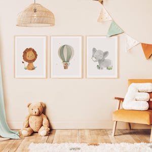 May include: Three framed watercolor prints featuring a lion, hot air balloon, and elephant. The lion is tan with a brown mane, the balloon is green and white, and the elephant is gray. The prints are displayed in a child's room with a teddy bear and a yellow chair.