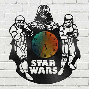May include: A black vinyl record clock featuring Star Wars characters Darth Vader, a Stormtrooper, and another Stormtrooper. The clock face is a rainbow gradient with black hands and black Roman numeral markings. The text "STAR WARS" is printed below the clock face.