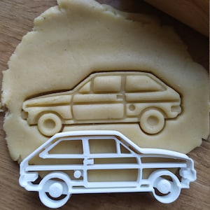 Może przedstawiać: Biała plastikowa foremka do ciasteczek w kształcie zabytkowego samochodu. Foremka leży na drewnianej powierzchni z kawałkiem ciasta z tyłu.