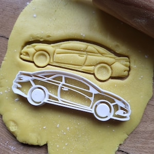Może przedstawiać: Wycinarka do ciastek w kształcie samochodu i ciasteczko w kształcie samochodu wciśnięte w żółte ciasto. Wycinarka do ciastek jest z białego plastiku, a wciśnięte ciasteczko jest jasnożółte. Drewniany wałek do ciasta znajduje się w tle.