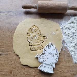 May include: A cookie cutter in the shape of a stylized bird is pressed into a circle of dough. A wooden rolling pin and a pile of flour are in the background. The cookie cutter is white and the dough is light brown.