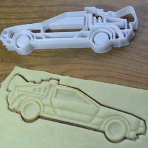 Könnte beinhalten: Ein weißer Plastik-Ausstecher in Form eines DeLorean-Zeitautos. Der Ausstecher liegt auf einer Holzoberfläche und im Hintergrund ist eine Keksmasse-Abdruck des DeLorean zu sehen.